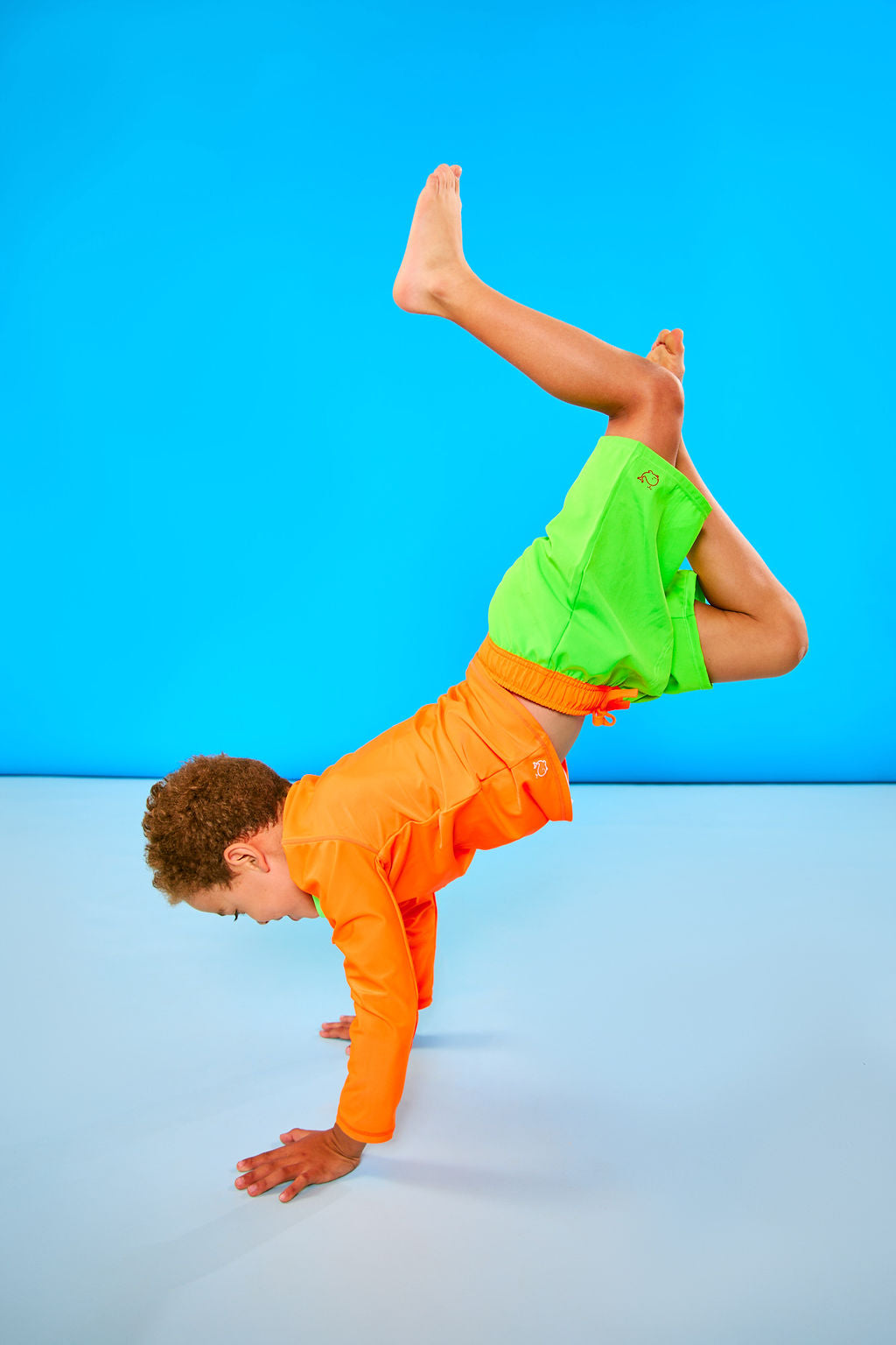 Child doing headstand wearing neon orange rash guard and neon green swim trunks