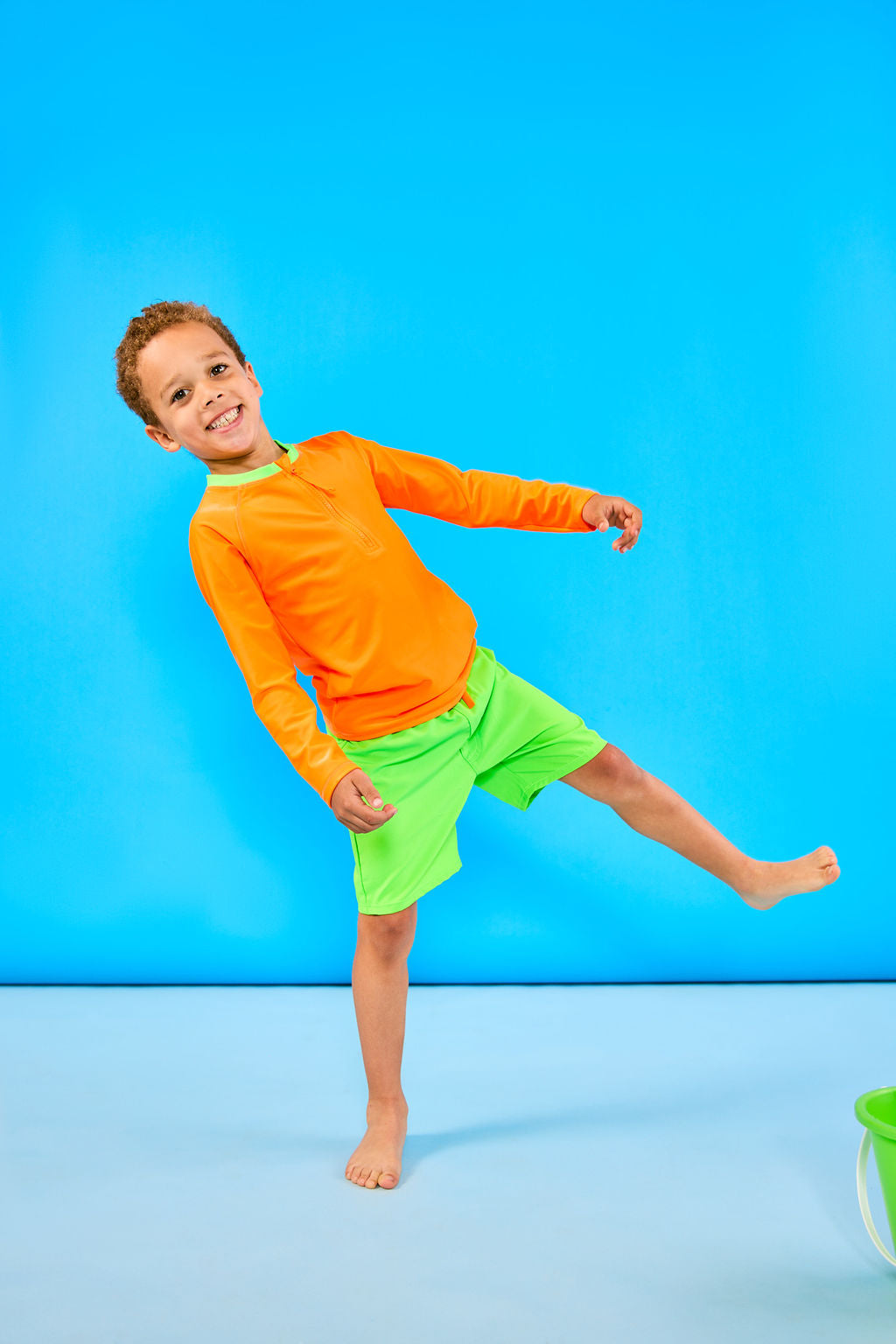 Child leaning sideways and smiling while wearing neon green swim trunks with a neon orange waistband and a neon orange rash guard with a neon green collar.