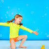 Child posing playfully wearing a neon yellow rash guard and neon orange euro swim shorts while surrounded by bubbles and next to a small rubber duck
